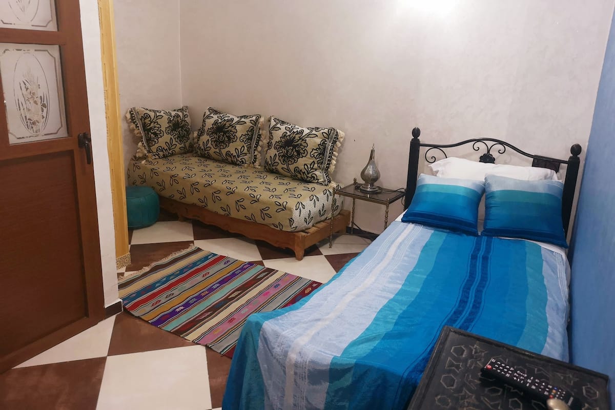 A bedroom with a bed, a sofa, a small table with a lamp, and a colorful rug.