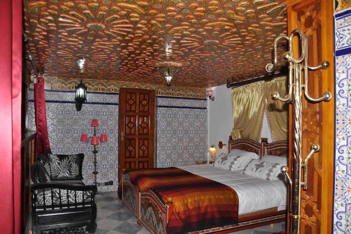 A traditional room with a patterned ceiling, a bed with a red and black bedspread, a wooden chair, and a red lamp.