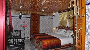 A traditional room with a patterned ceiling, a bed with a red and black bedspread, a wooden chair, and a red lamp. - Riad Royal (Meknes)