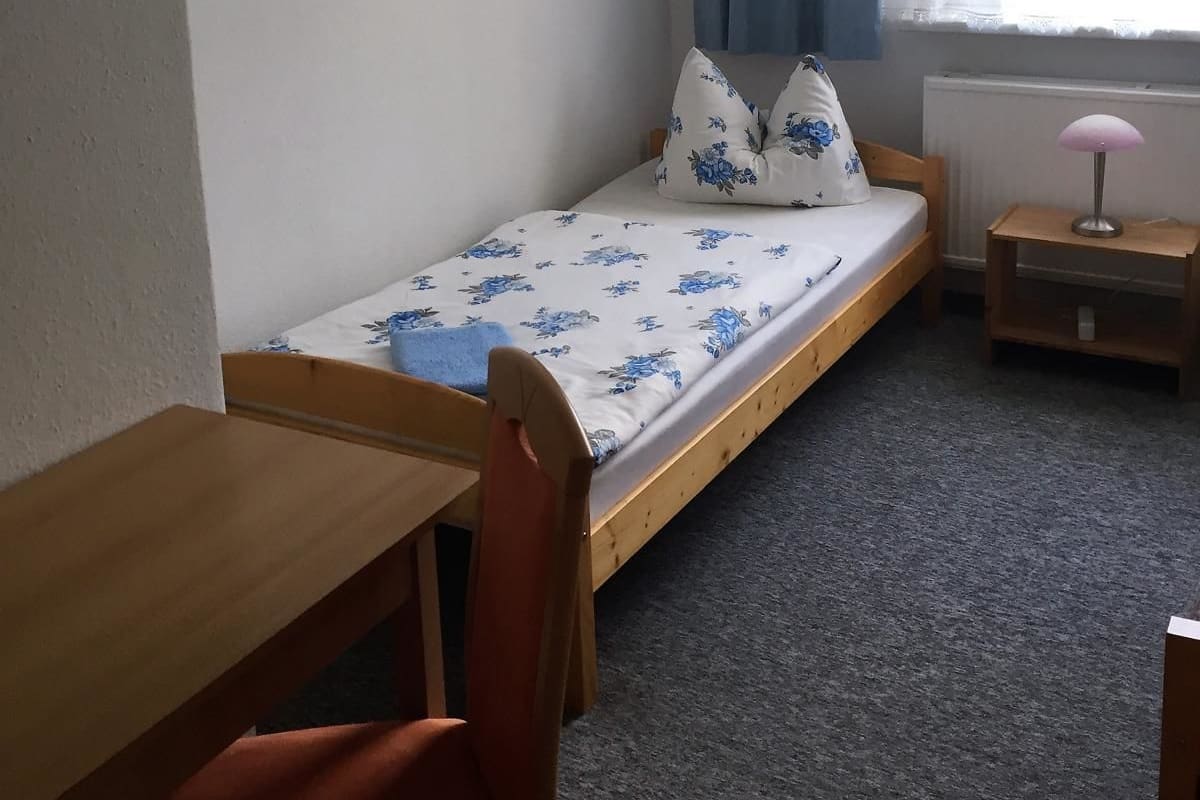 A small, single-bed room with a wooden desk and chair, a bedside table with a lamp, and a window with curtains.