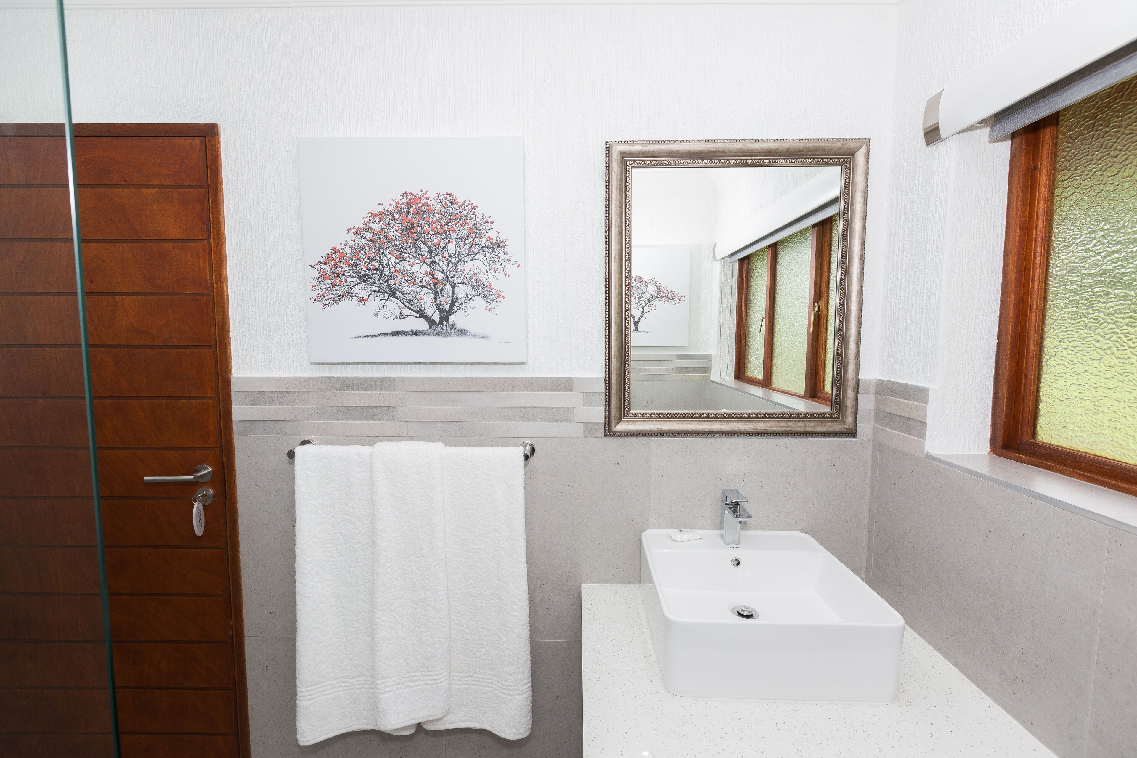 A bathroom with a wooden door, a white sink, a mirror, and a framed artwork of a tree.