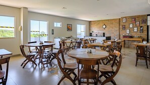 A spacious dining area with multiple tables and chairs, a kitchen area with a brick wall, and large windows offering a view of greenery. - OYO Tropical Confort Hotel, Brasilia (São Sebastião)