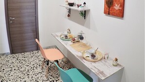 Une pièce avec une porte en bois, un fauteuil de couleur turquoise, une table blanche avec un plateau en verre et des étagères murales ornées d’œuvres d’art et de petites figurines.