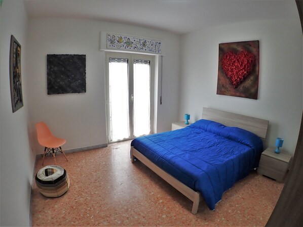 Un dormitorio con cama, una silla, una mesita de noche, una obra de arte en la pared y una puerta con un panel decorativo.