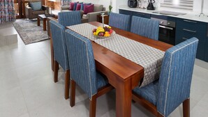 A dining area with a wooden table and blue upholstered chairs, a fruit bowl on the table, and a kitchen in the background. - Kruger Park Lodge Unit 608B (Mbombela)