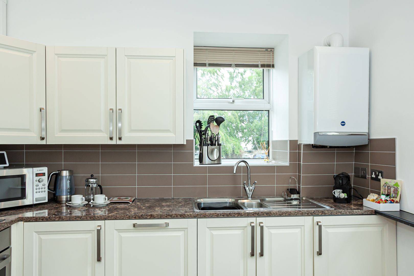 A modern kitchen with white cabinets, a built-in microwave, a coffee maker, and a window with a view of trees.