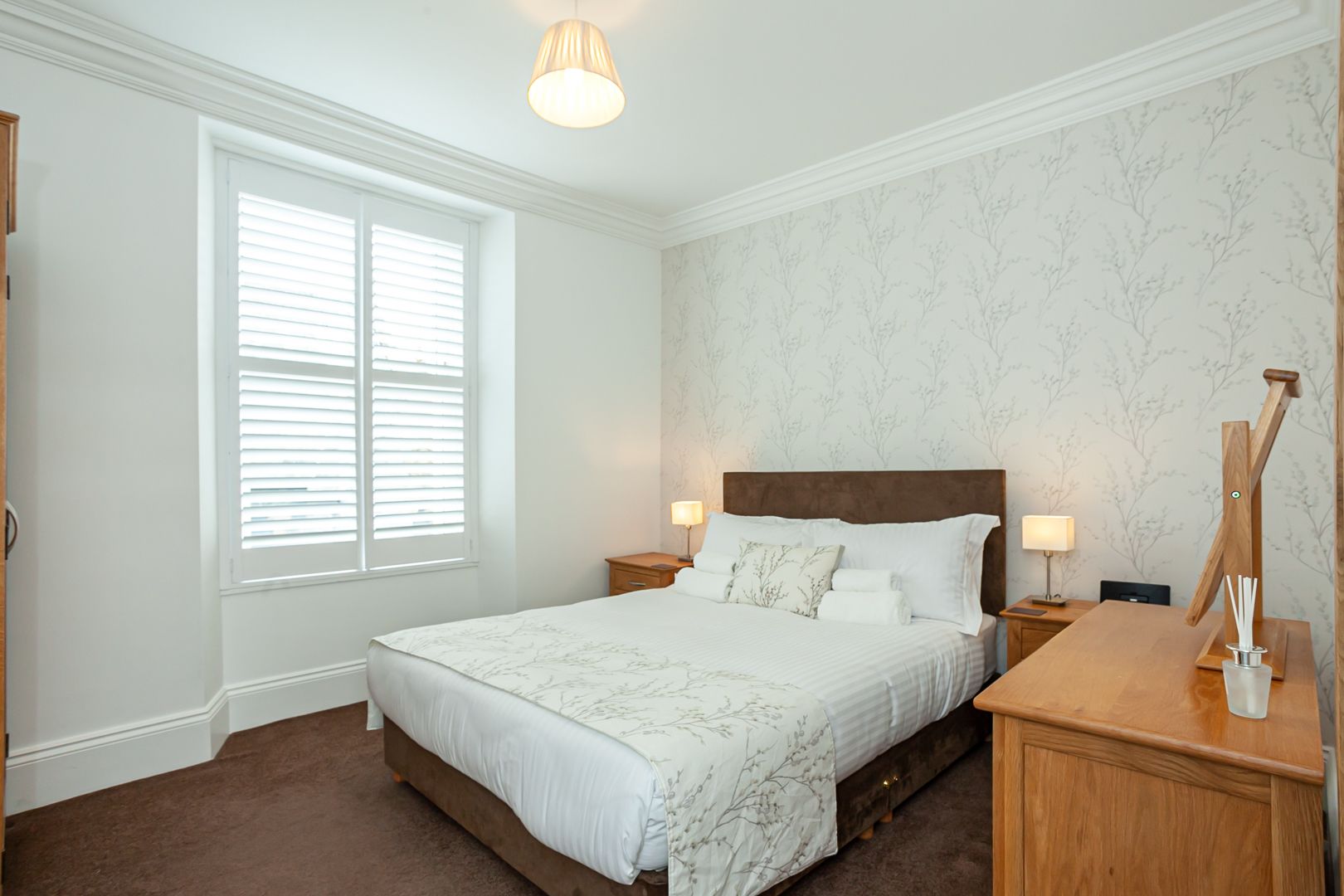 A bedroom with a large bed, two bedside tables, a window with blinds, and a wall with a subtle pattern.