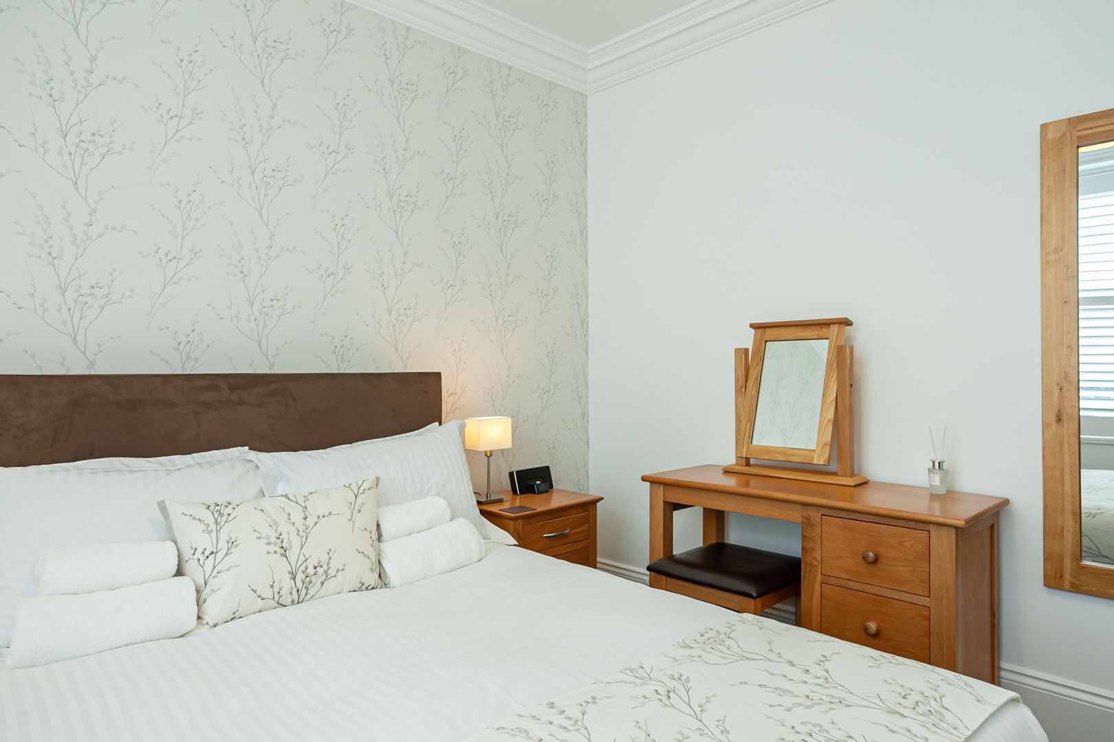 A neatly made bed with a wooden headboard, a nightstand with a lamp, a wooden dresser with a mirror, and a wall with a floral wallpaper pattern.