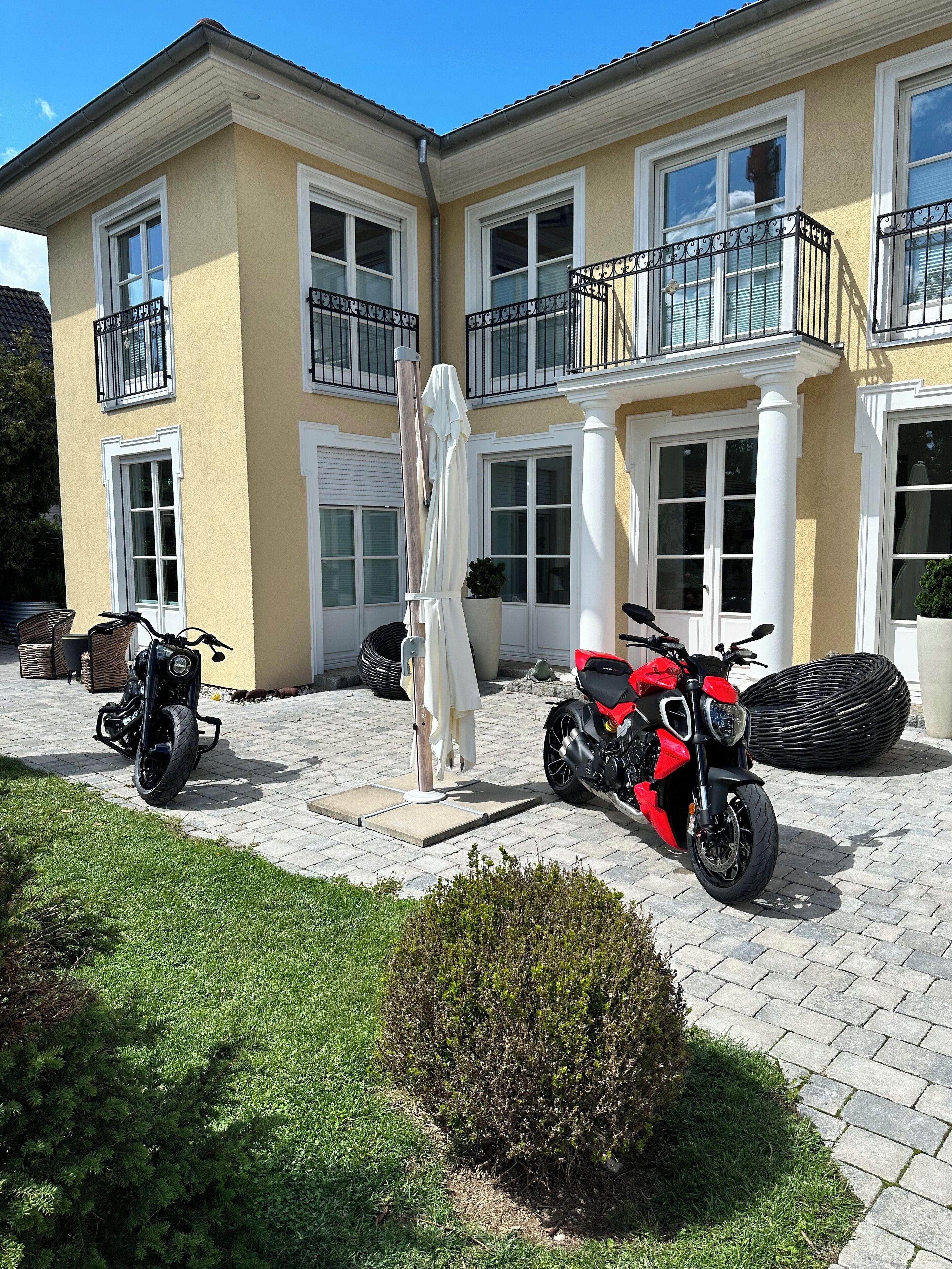 Una casa de dos pisos con un camino de entrada pavimentado, dos motocicletas estacionadas afuera y un paraguas blanco.