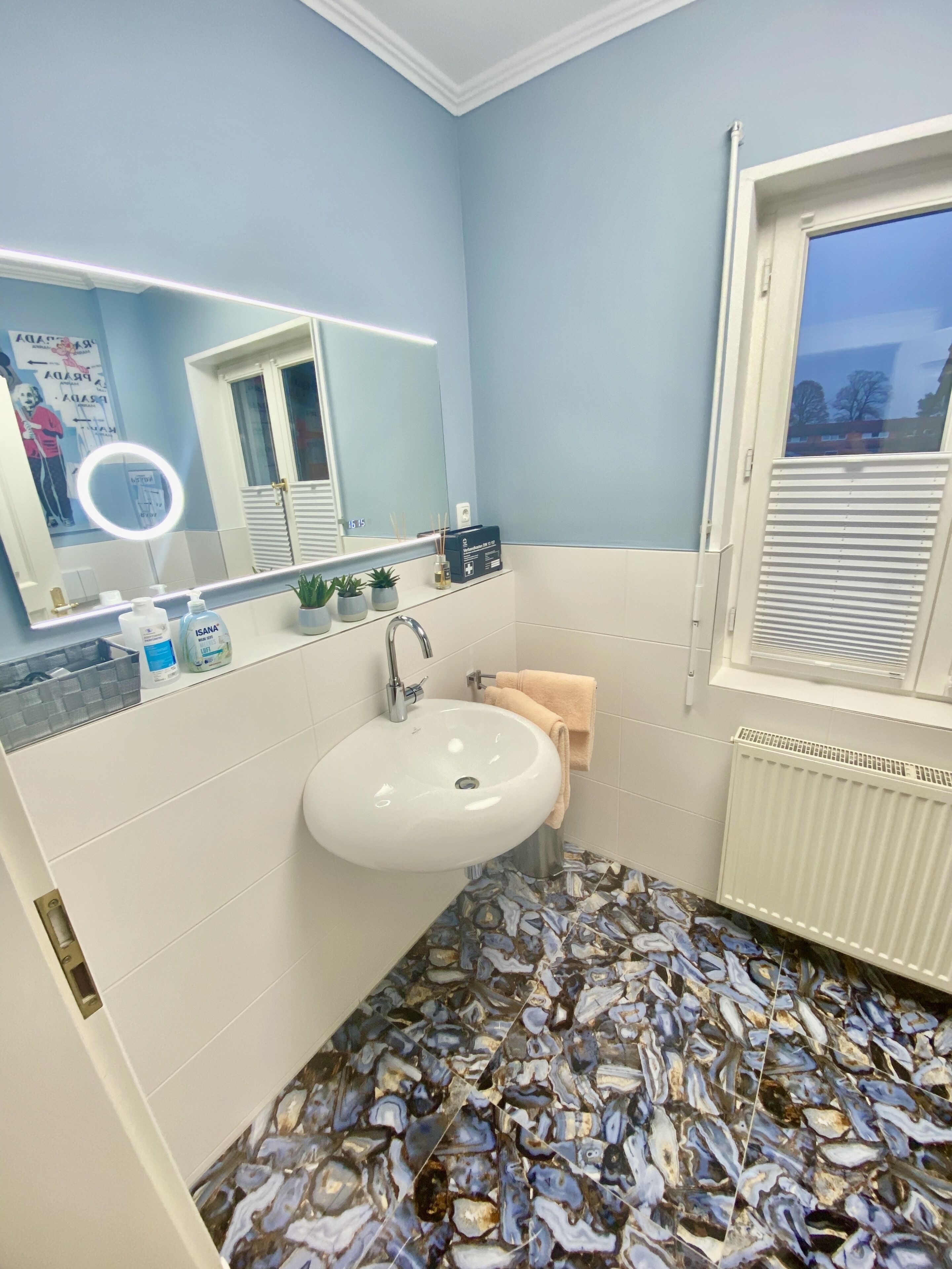 Un baño con un lavamanos blanco, un espejo con luz, una ventana y un suelo estampado.