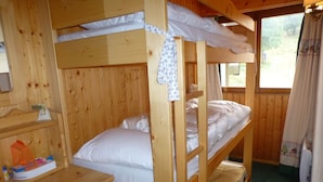 A bunk bed room with wooden walls and a window with curtains. There is a small wooden dresser with a mirror and a shelf with items on it.