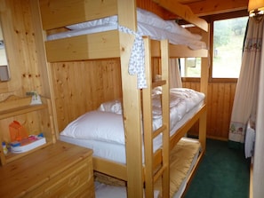 Quarto com beliche, paredes de madeira e janela com cortinas. Há uma pequena cômoda de madeira com espelho e uma prateleira com objetos.