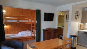 Una habitación compacta con una cama litera, una mesa de comedor de madera con bancos, un sofá gris, una pequeña cocina con un fregadero y una estufa, y un televisor montado en la pared.