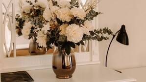 A white desk with a vase of white flowers, a framed picture on the wall, and a TV mounted above. - Golden Hotel (Pristina)