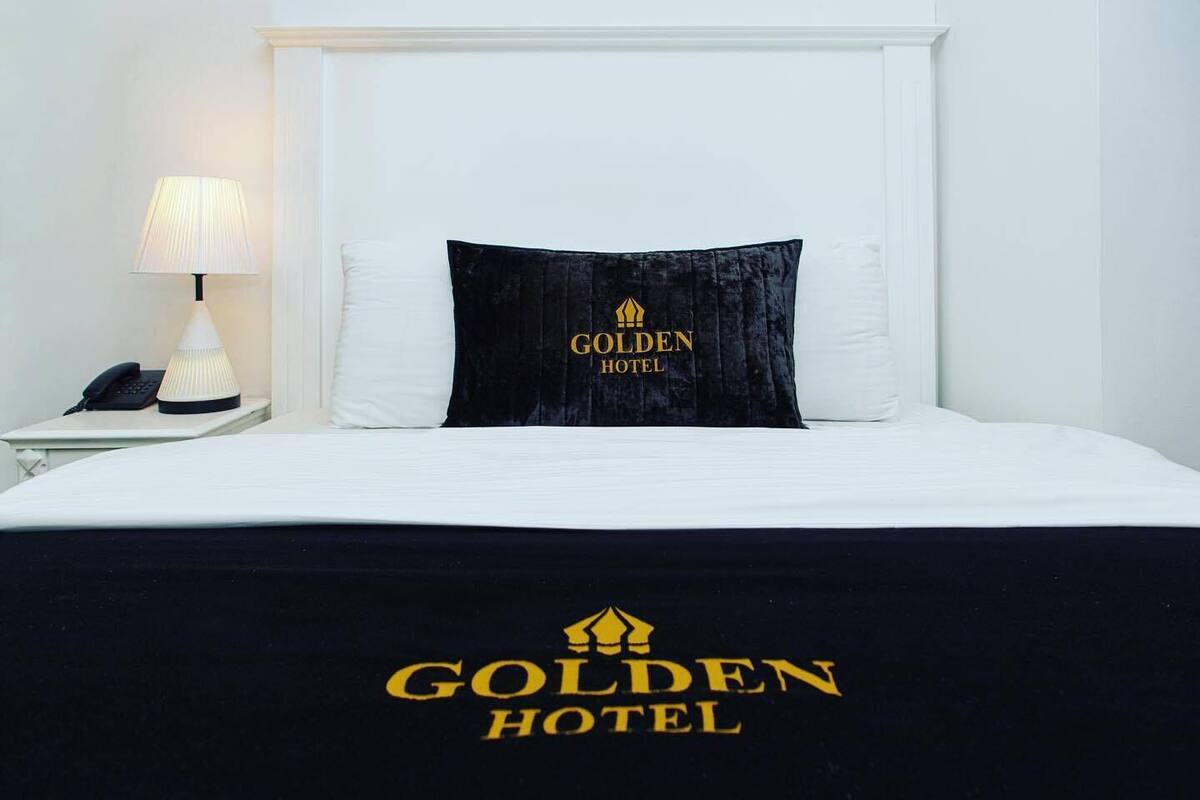 A hotel room with a bed, a nightstand with a lamp, and a pillow with the hotel's logo.