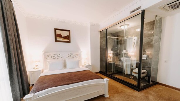 A bedroom with a bed, a nightstand, a bedside table, a glass-enclosed bathroom, and a painting on the wall. - Golden Hotel (Pristina)