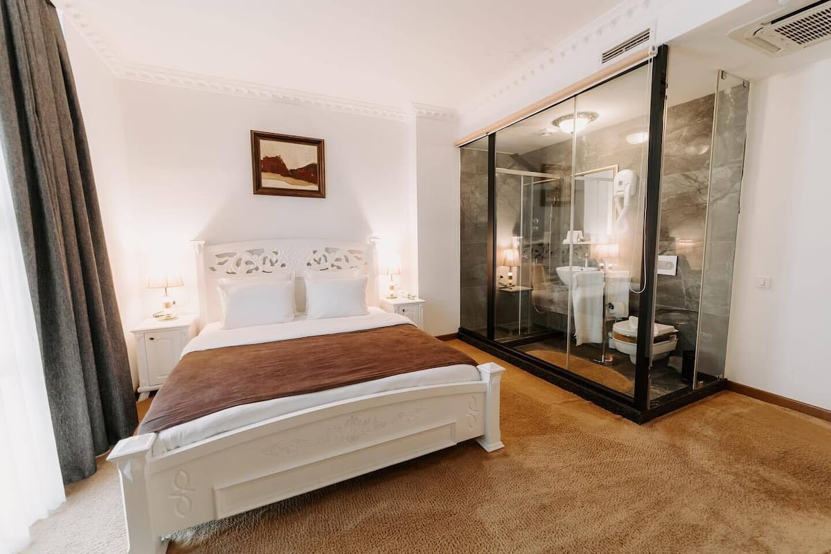 A bedroom with a bed, a nightstand, a bedside table, a glass-enclosed bathroom, and a painting on the wall.