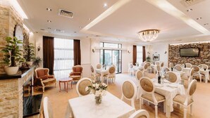 A spacious hotel dining area with multiple tables and chairs, a chandelier, and a stone wall with a television. - Golden Hotel (Pristina)