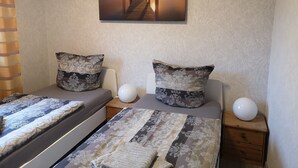 Een slaapkamer met een bed, twee kussens, een nachtkastje met een lamp en een wanddecoratie van een pier bij zonsondergang.
