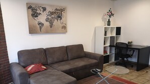 Een woonkamer met een grijze bank, een klein voetenbankje, een glazen salontafel, een wereldkaart als kunstwerk, een witte boekenkast en een zwarte bureaustoel.
