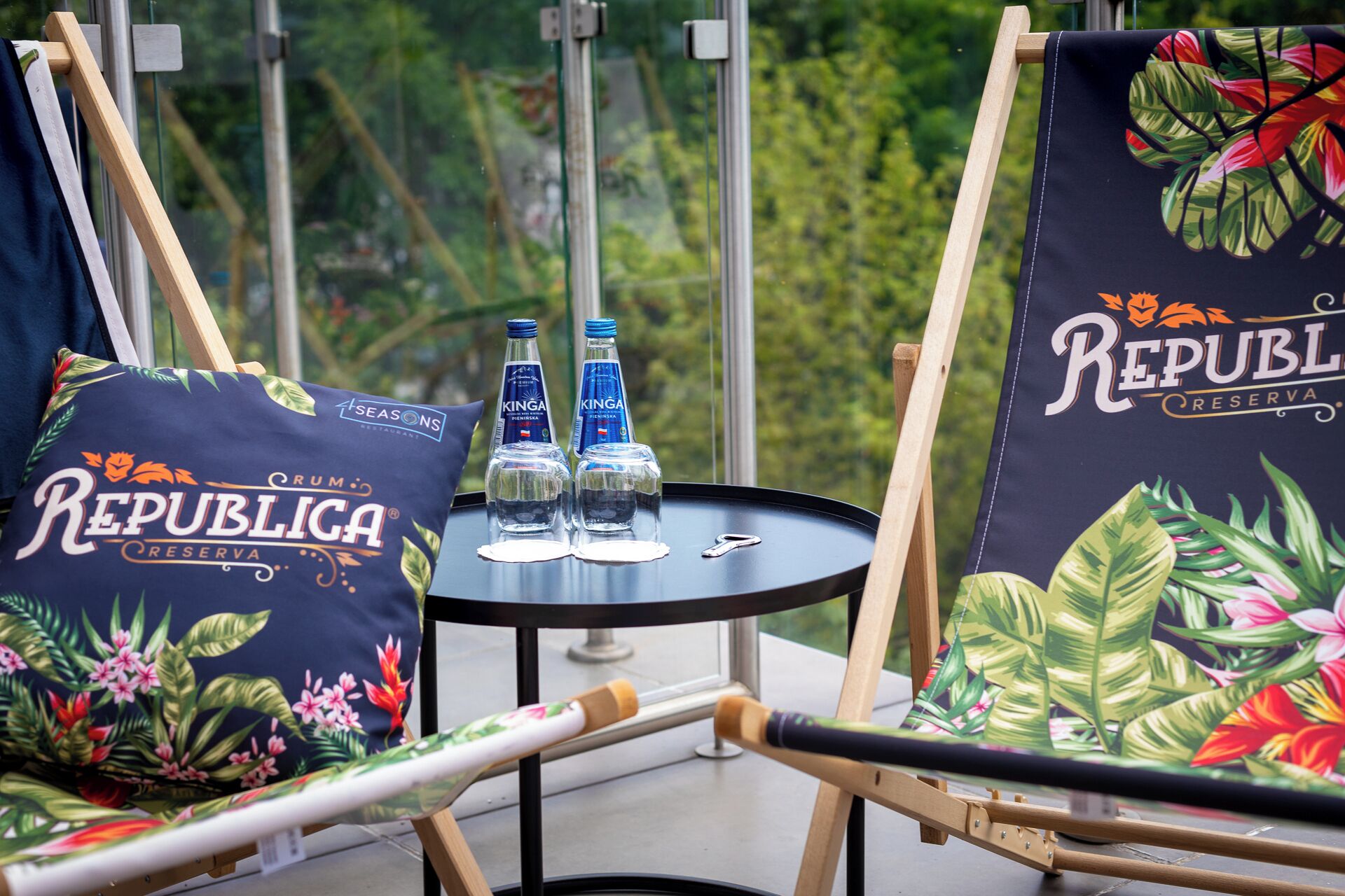 A balcony with two chairs, each with a pillow featuring 'Republica Reserva' branding, a small table with two bottles of water, and a view of greenery through a glass door.