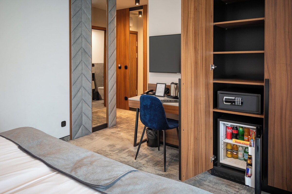 A compact hotel room with a bed, a desk, a chair, a minibar, and a television.