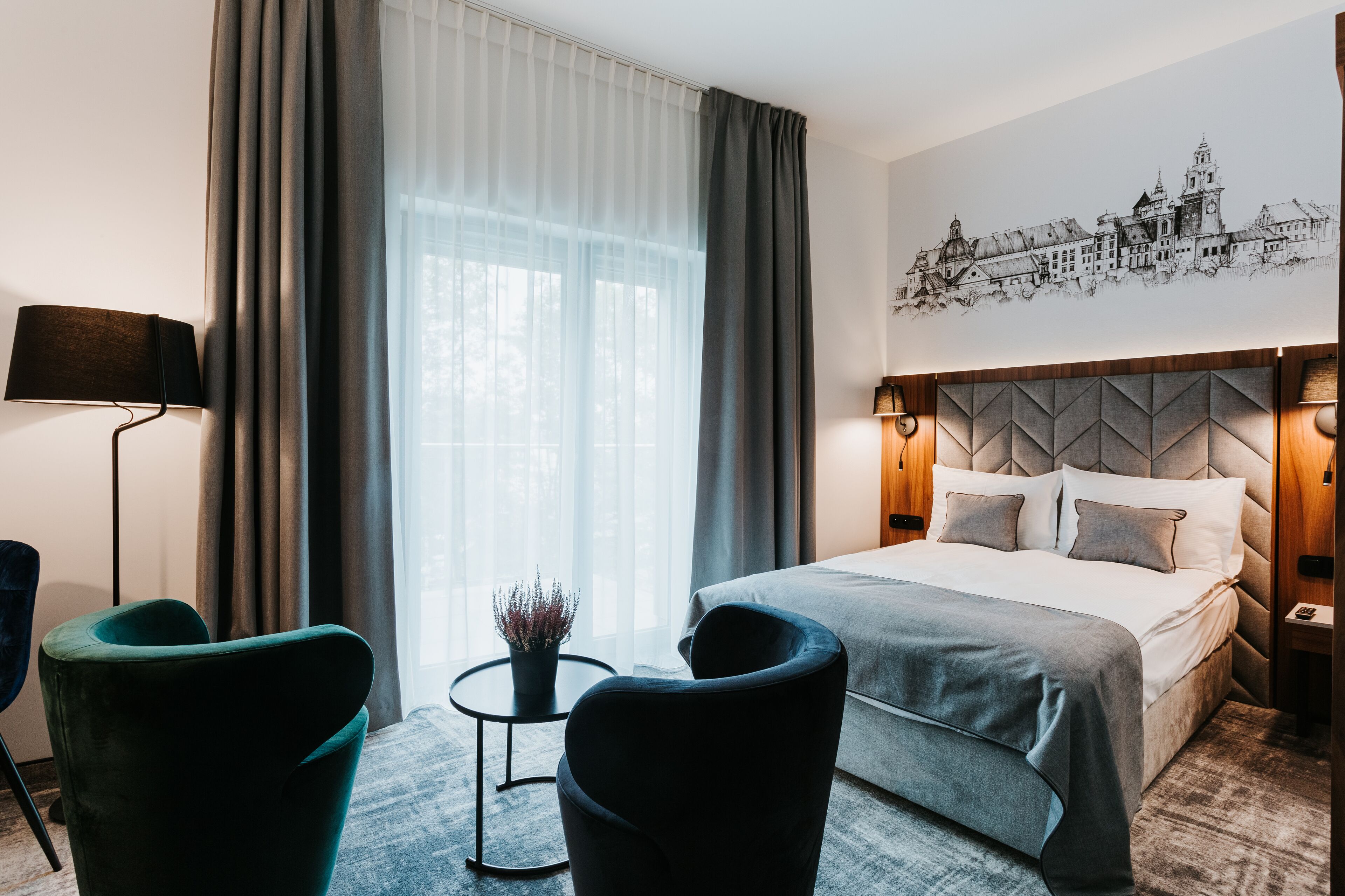 Une chambre d’hôtel moderne avec un grand lit, deux fauteuils, une petite table avec une plante et une œuvre d’art représentant un paysage urbain accrochée au mur.
