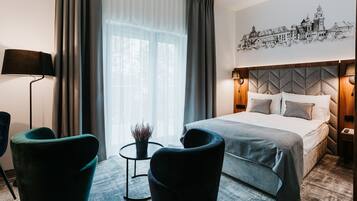 Une chambre d’hôtel moderne avec un grand lit, deux fauteuils, une petite table avec une plante et une œuvre d’art représentant un paysage urbain accrochée au mur.