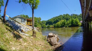 Een huis aan de rivier met een houten terras en een grote boom ervoor.