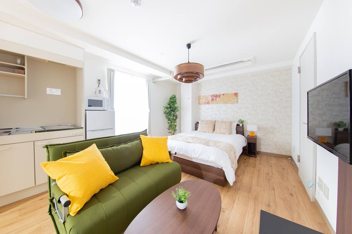 Un espace de vie compact comprenant un canapé vert, un lit avec une literie blanche, une table basse en bois, un téléviseur et un coin cuisine.