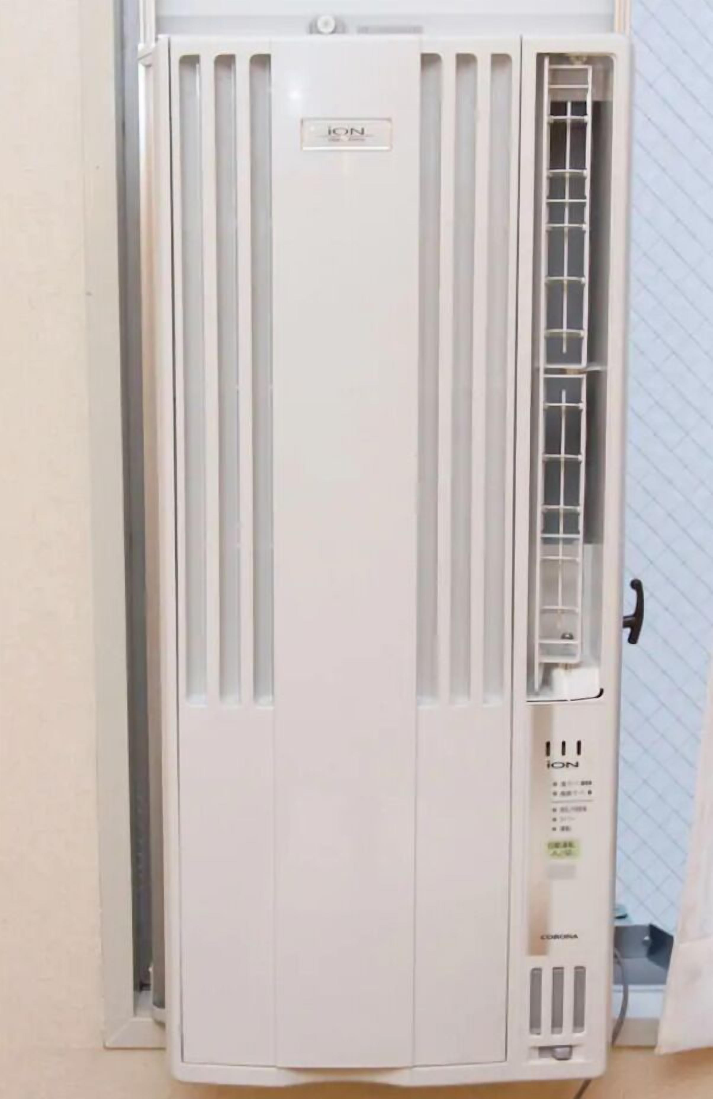 A white portable air conditioner with a control panel on the right side, placed against a wall with a window.