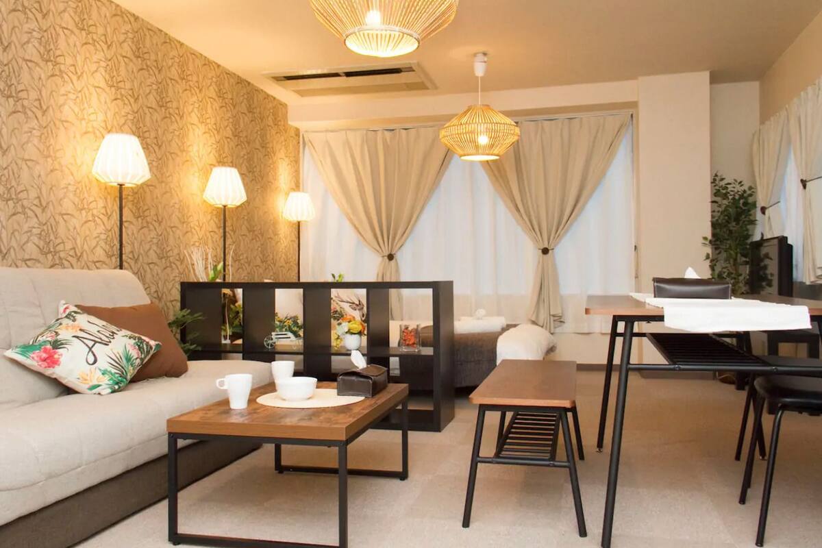 Un salon moderne avec un canapé, une table basse et un coin repas. On y trouve deux lampes, une plante et une vue sur l’extérieur grâce à de grandes fenêtres.