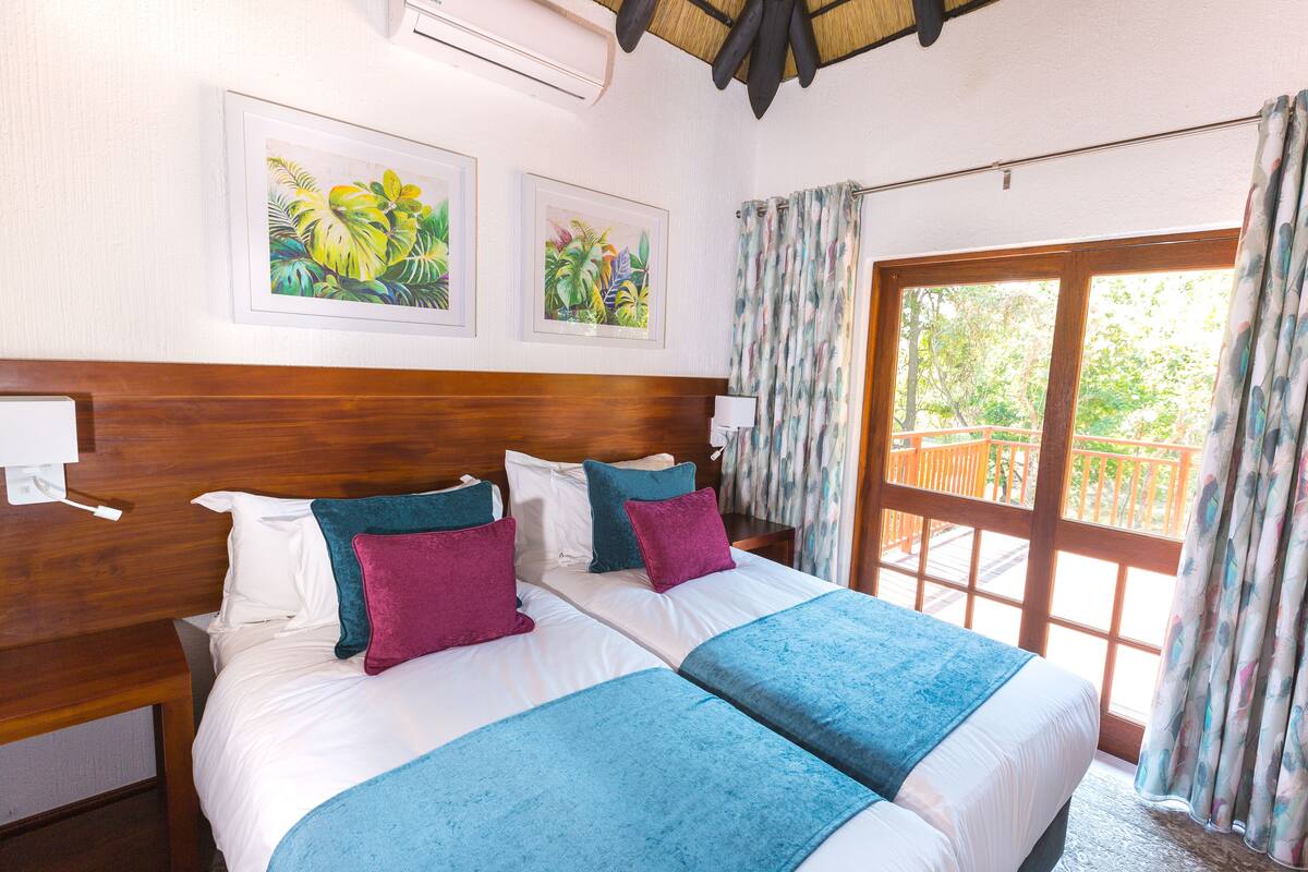 A double bed with two pillows, a nightstand, a wooden headboard, and two framed tropical paintings on the wall.