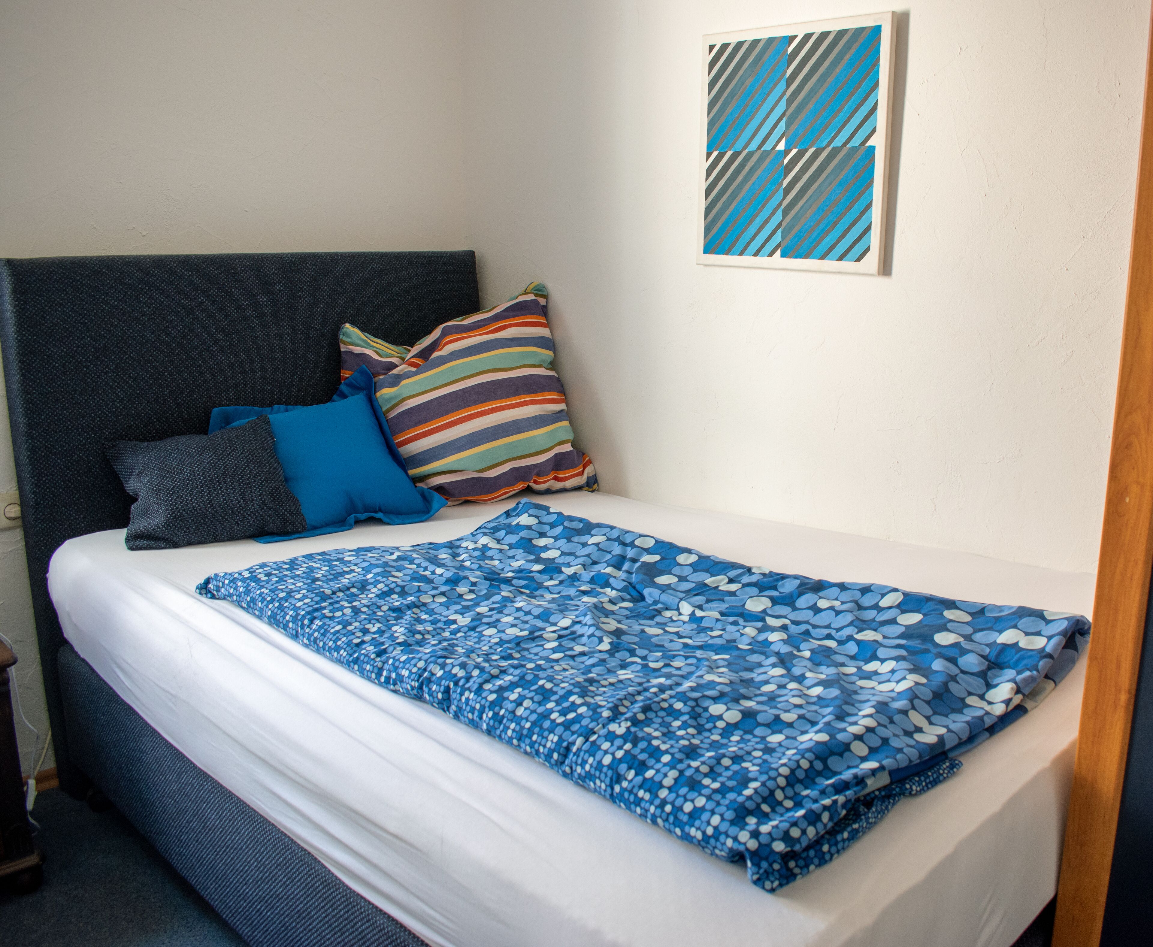 A bed with a blue patterned blanket, a striped pillow, and a blue pillow.