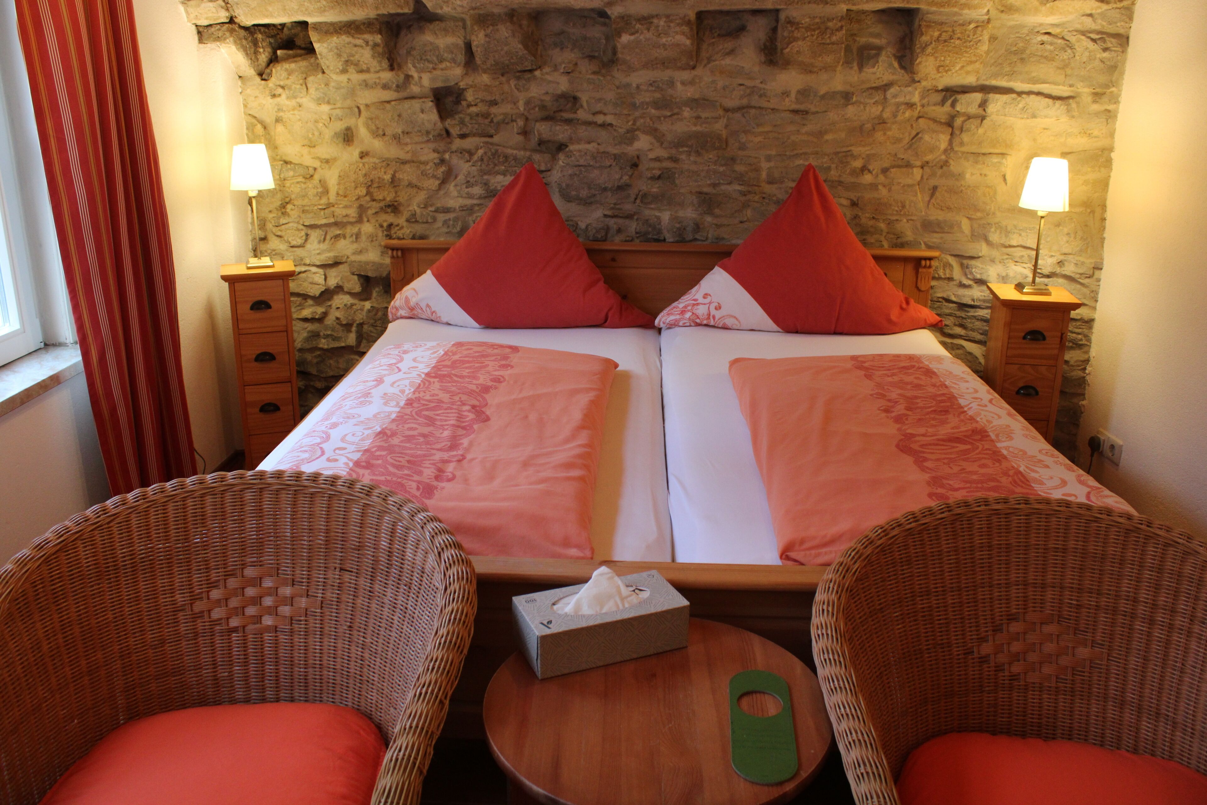 A double bed with red and white linens, two nightstands with lamps, wicker chairs, and a small table with a tissue box.