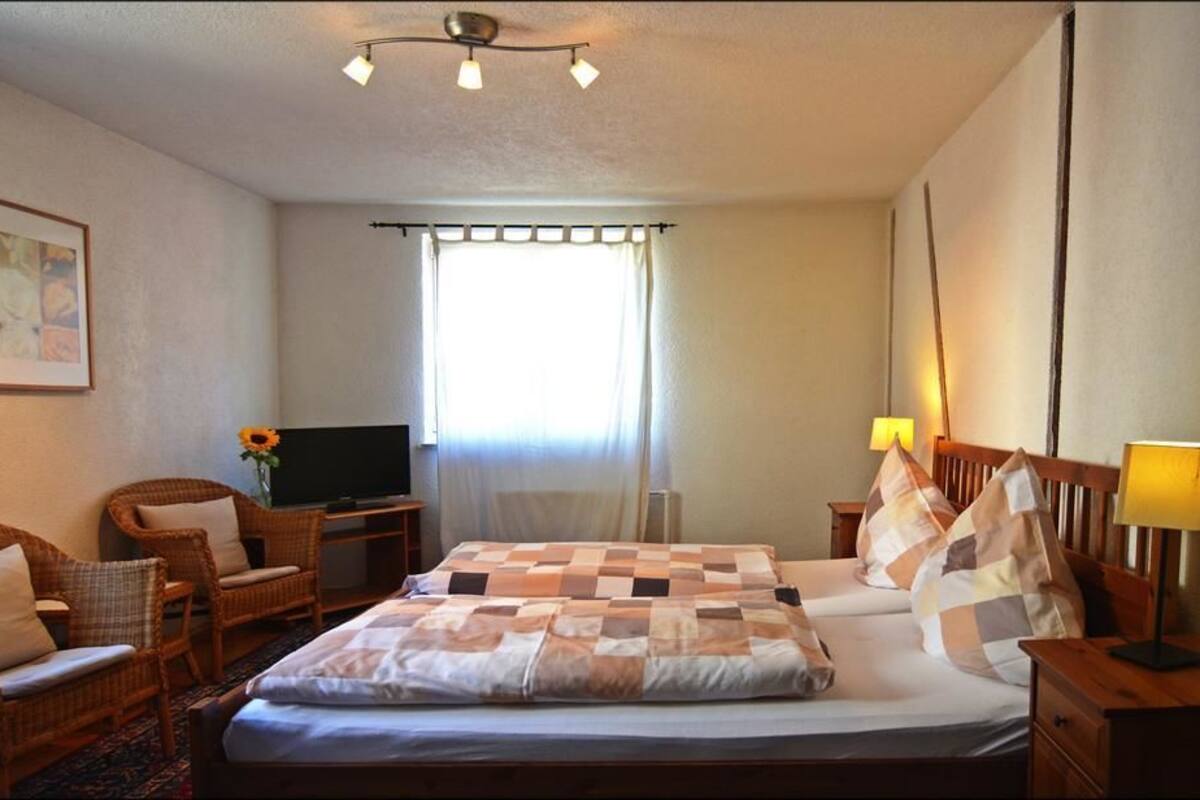 Hotellihuone, jossa on sänky, kaksi tuolia, televisio, lamppu ja ikkuna, jossa on verhot.