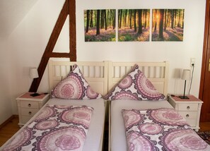 A bedroom with two beds, each with patterned bedding, a wooden headboard, bedside tables with lamps, and a wall with three framed pictures.
