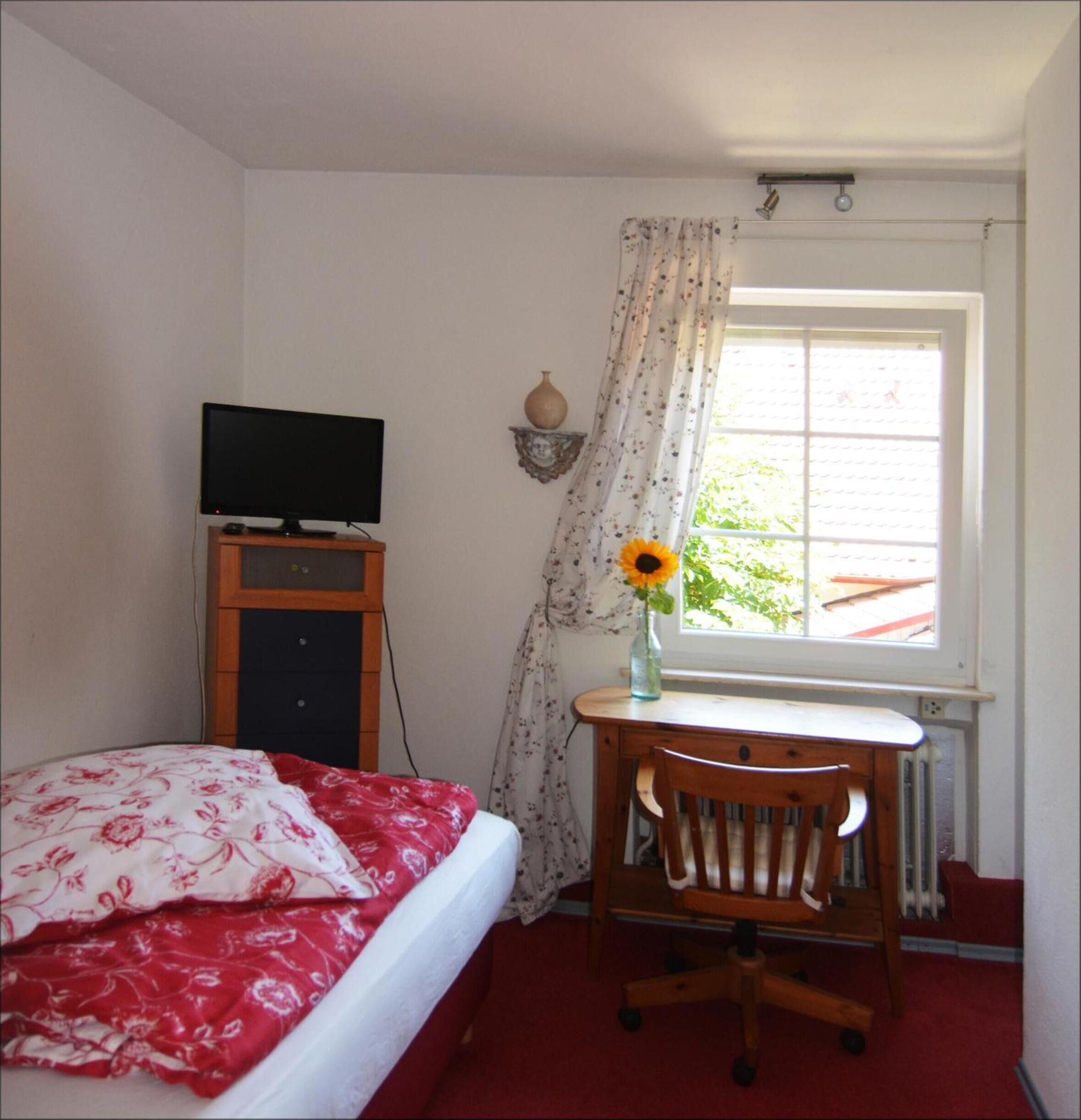 Un dormitorio con cama, escritorio con silla, televisor sobre un mueble bajo, ventana con cortinas y radiador.