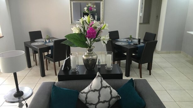 A living room with a grey sofa, teal cushions, a coffee table with a vase of flowers, and a dining area with a table and chairs. - The Grand Orchid Guesthouse (Durban North)