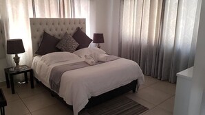 A neatly made bed with a tufted headboard, two bedside lamps, and a patterned throw blanket. - The Grand Orchid Guesthouse (Durban North)