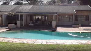 A swimming pool with a tiled deck, a covered patio area with seating, and a building with a tiled roof. - The Grand Orchid Guesthouse (Durban North)