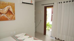 A hotel room with a bed, an air conditioner, a painting on the wall, and a view of greenery outside. - Suites Solanas de Porto - Porto de Galinhas (Ipojuca)