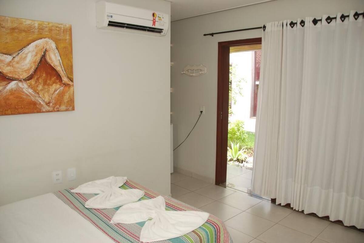 A hotel room with a bed, an air conditioner, a painting on the wall, and a view of greenery outside.