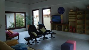 A spacious living room with a large window, a television, and a bookshelf. - Suites Solanas de Porto - Porto de Galinhas (Ipojuca)