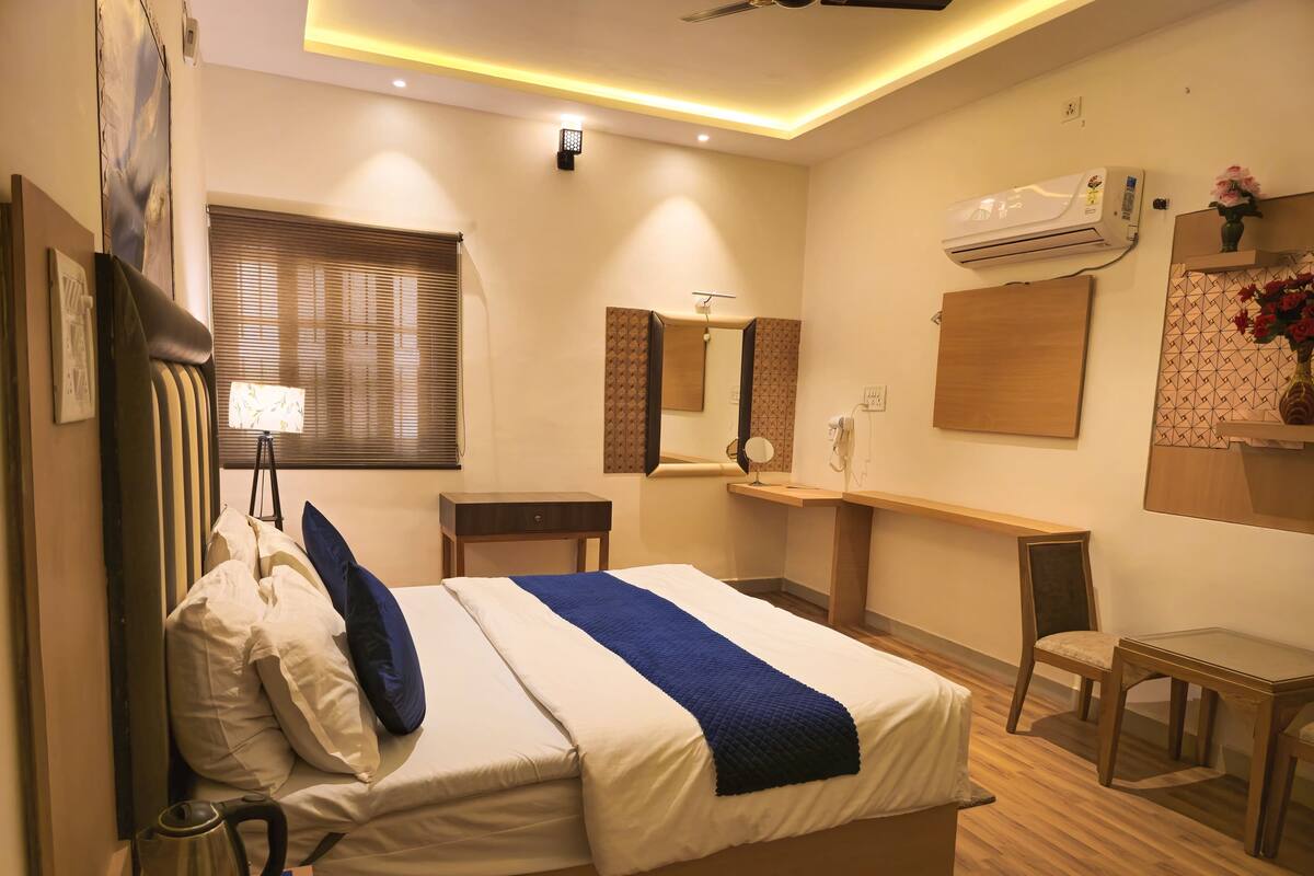 A hotel room with a bed, bedside table, lamp, mirror, air conditioning unit, and a small desk with a chair.