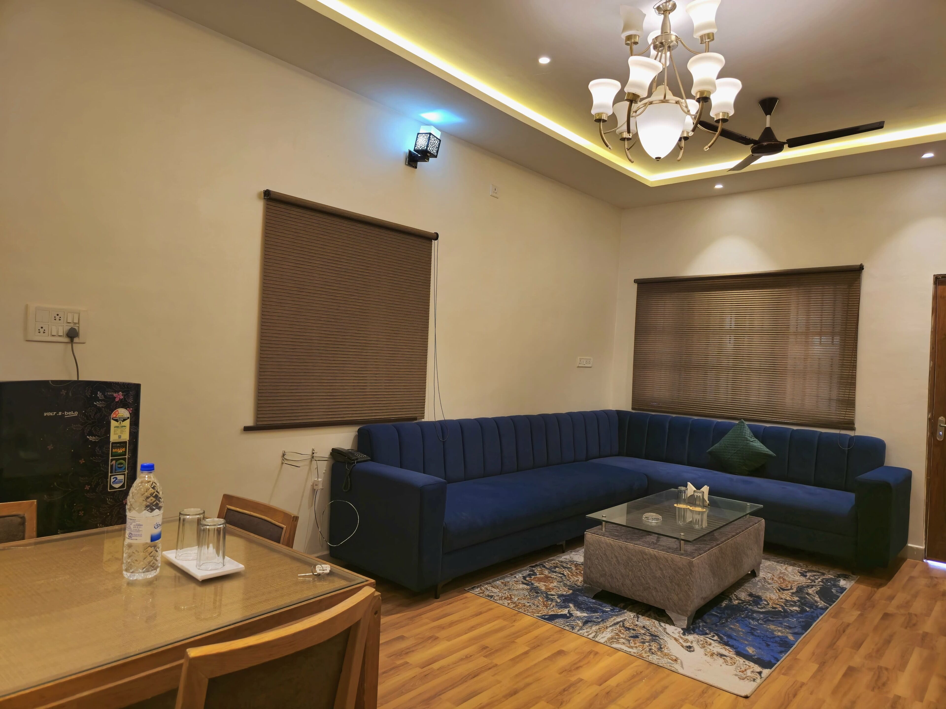 A living room with a blue sofa, a glass coffee table, a wooden dining table with chairs, and a patterned rug.