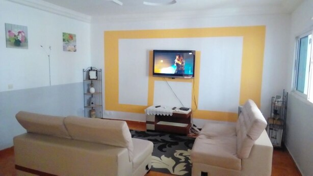 A living room with two beige sofas, a flat-screen TV mounted on a yellow wall, and a small table with a white tablecloth. - Rental Studio Floor Villa (Abidjan)