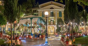 A well-lit courtyard with a covered seating area, potted plants, and a large building in the background. - Kathmandu Guest House by KGH Group (Kathmandu)