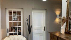Ein Zimmer mit weißer Tür, einem Korbstuhl, einem Holzschrank und einem Spiegel mit Zierrahmen.