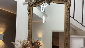 Ein großer, aufwendig verzierter Spiegel mit goldener Rahmung, aufgestellt auf einem Holzboden neben einem weißen Strauß getrockneter Blumen.